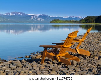 Two empty wooden Adirondack chairs or Muskoka deckchairs on stony shore overlooking scenic calm Lake Laberge, Yukon Territory, Canada, with snowcapped mountains in the distance