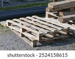 Two empty wooden pallets stand on the edge of a construction site on rubble and are illuminated by the sun from behind on the left. Next to them are pallets with lumber on them and black district heat