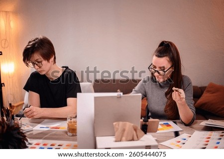 Two engaged artists at work, one examining her brush stroke, the other evaluating color options, showcasing their intense focus in a creative studio space