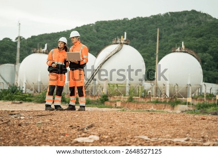 Two engineer workers review data on laptop, team discusses plans at energy plant, Petrochemicals factory, Storage tanks, construction site survey, Industrial site operation