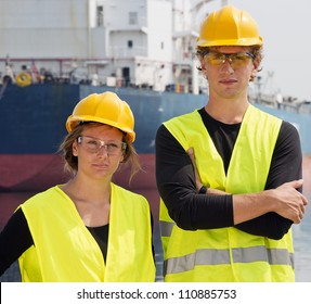 Two Engineering Students Posing In Front Of A Huge Freight Vessel During Their Technological Internship In A Harbor