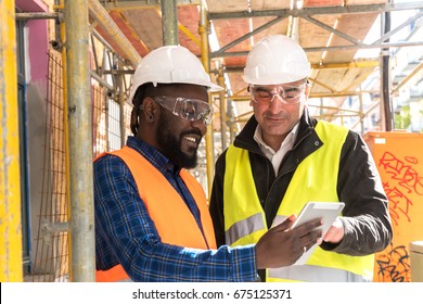 Two Engineers, An African American And A Caucasian, Wearing Orange And Yellow Safety Jackets And Helmets, Working With Tablet Computer Among Scaffolding On Construction Site