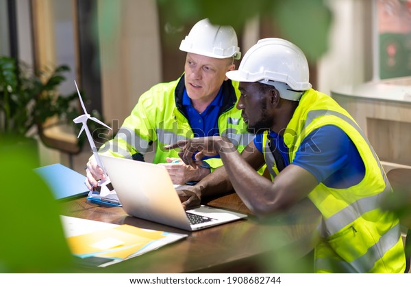Two\
engineers African american engineer and caucasian electrician\
wearing white hard hat working on laptop computer at workplace\
office. clean and green alternative energy\
concept.