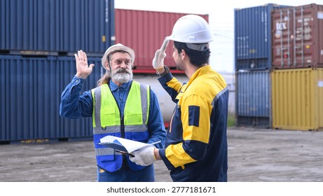 Two engineers celebrate project completion in a container yard. They high-five, smiling, wearing hard hats and safety vests. One holds blueprints, gesturing with open hands