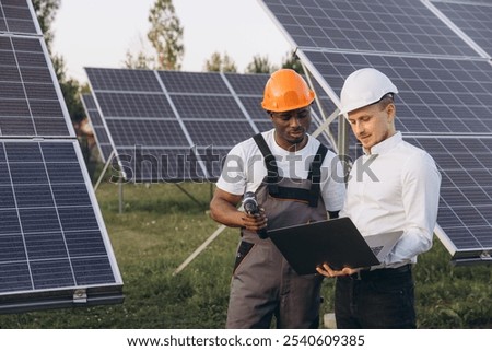 Two engineers collaborate onsite amidst a solar panel array, evaluating data on a laptop. They wear safety helmets, emphasizing renewable energy solutions and teamwork in the green energy sector.