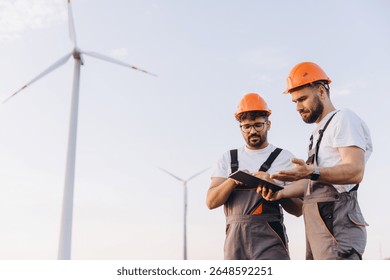 Two engineers collaborating on a tablet while working at a wind turbine farm, contributing to sustainable energy production and innovation