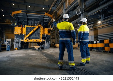 Two engineers control repair of dump car in garage of factory. Industry big yellow mining truck maintenance.