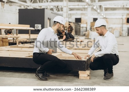 Two engineers crouching and inspecting modular building components in a factory, ensuring quality and precision in prefabricated construction