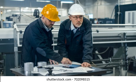 Two Engineers Discuss A Blueprint While Checking Information On A Tablet Computer In A Factory.