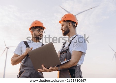 Two engineers discussing using laptop in a wind turbine farm, working on renewable energy development