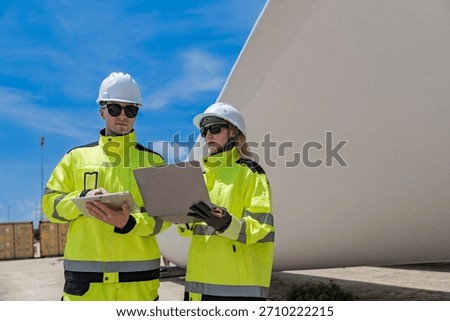 Two engineers in high visibility gear use a laptop to work in front of a large wind turbine blade at a construction site. the integration of technology in renewable energy and wind power projects.