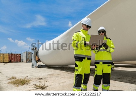 Two engineers in high visibility gear use a laptop to work in front of a large wind turbine blade at a construction site. the integration of technology in renewable energy and wind power projects.
