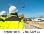 Two engineers in high visibility jackets and helmets observing a drone flying over a construction site, with machinery and modular buildings, highlighting technology and site monitoring.