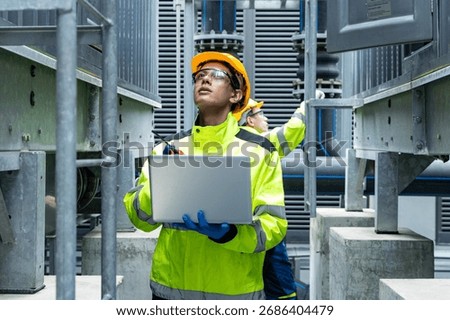 Two engineers in high-visibility gear and helmets examine a control panel on industrial HVAC equipment, using a walkie-talkie to coordinate.