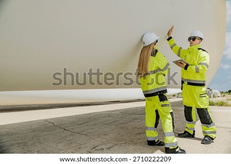 Two engineers in high-visibility gear inspect a large wind turbine blade at a construction or installation site. renewable energy, wind power, and the detailed work involved in energy projects.