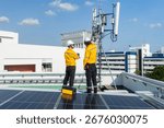 Two engineers in high-visibility jackets and helmets point and discuss work near a rooftop telecommunications antenna in an urban area.