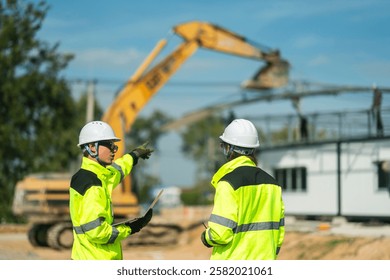Two engineers in high-visibility jackets and helmets reviewing plans on a laptop at a construction site with an excavator in the background, representing safety, precision, and technological planning.
