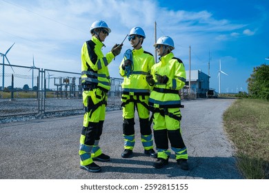 Two engineers in high-visibility jackets and helmets discuss project plans at a wind farm, using handheld devices and protective equipment.