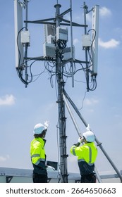 Two engineers in high-visibility jackets and helmets point and discuss work near a rooftop telecommunications antenna in an urban area.