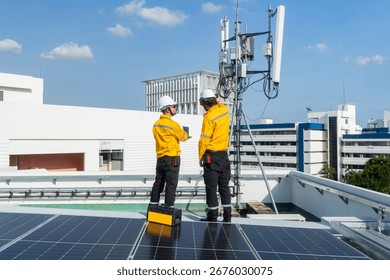 Two engineers in high-visibility jackets and helmets point and discuss work near a rooftop telecommunications antenna in an urban area.