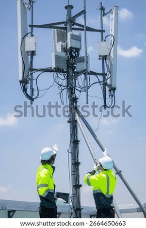 Two engineers in high-visibility jackets and helmets point and discuss work near a rooftop telecommunications antenna in an urban area.