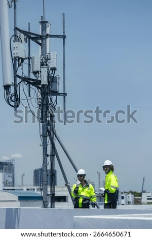 Two engineers in high-visibility jackets and helmets point and discuss work near a rooftop telecommunications antenna in an urban area.