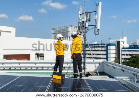 Two engineers in high-visibility jackets and helmets point and discuss work near a rooftop telecommunications antenna in an urban area.