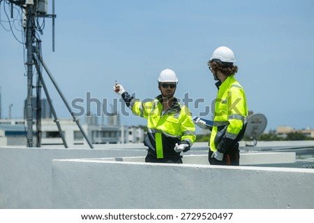 Two engineers in high-visibility jackets and helmets point and discuss work near a rooftop telecommunications antenna in an urban area.