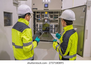 Two engineers inspect an electrical control panel in a factory, recording data on a tablet and clipboard. Professional maintenance, power distribution, and industrial safety inspection concept.