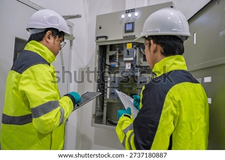 Two engineers inspect an electrical control panel in a factory, recording data on a tablet and clipboard. Professional maintenance, power distribution, and industrial safety inspection concept.