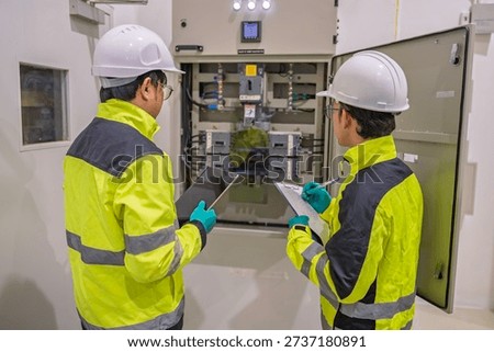Two engineers inspect an electrical control panel in a factory, recording data on a tablet and clipboard. Professional maintenance, power distribution, and industrial safety inspection concept.