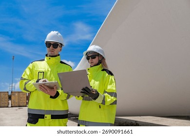 Two engineers inspect a massive wind turbine blade at a construction site. The image showcases the scale of wind power projects and the detailed work involved in renewable energy development.