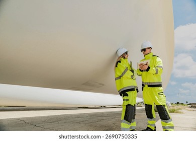 Two engineers inspect a massive wind turbine blade at a construction site. The image showcases the scale of wind power projects and the detailed work involved in renewable energy development.