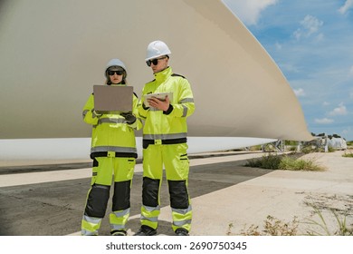 Two engineers inspect a massive wind turbine blade at a construction site. The image showcases the scale of wind power projects and the detailed work involved in renewable energy development.