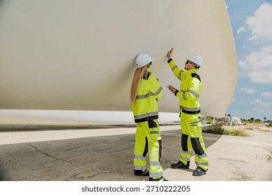 Two engineers inspect a massive wind turbine blade at a construction site. The image showcases the scale of wind power projects and the detailed work involved in renewable energy development.