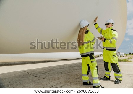 Two engineers inspect a massive wind turbine blade at a construction site. The image showcases the scale of wind power projects and the detailed work involved in renewable energy development.