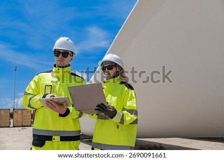 Two engineers inspect a massive wind turbine blade at a construction site. The image showcases the scale of wind power projects and the detailed work involved in renewable energy development.