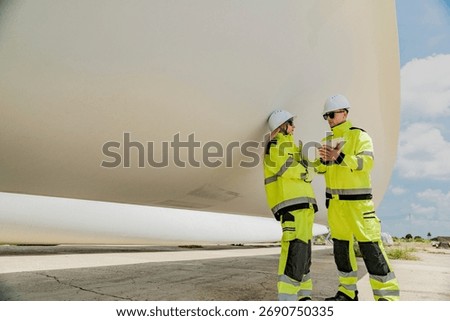 Two engineers inspect a massive wind turbine blade at a construction site. The image showcases the scale of wind power projects and the detailed work involved in renewable energy development.