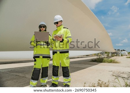 Two engineers inspect a massive wind turbine blade at a construction site. The image showcases the scale of wind power projects and the detailed work involved in renewable energy development.
