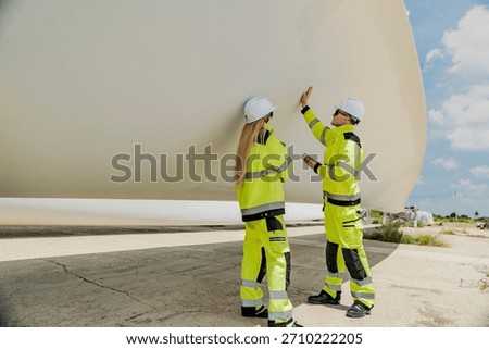 Two engineers inspect a massive wind turbine blade at a construction site. The image showcases the scale of wind power projects and the detailed work involved in renewable energy development.