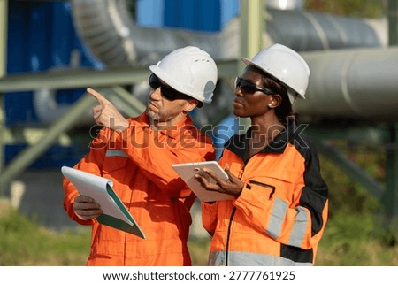 Two engineers in orange safety gear and helmets discuss a project outdoors, using a tablet and clipboard at an industrial site.