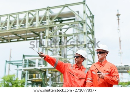 Two engineers in protective suits communicate via radio while pointing toward structural components of industrial plant, coordinating field inspection, safety assessment, and operational monitoring.