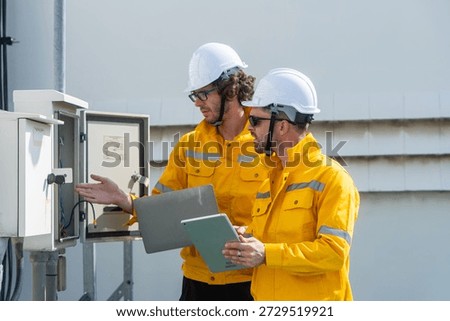 Two engineers in safety helmets and high-visibility clothing check rooftop electrical and communication equipment using tools and a digital tablet.