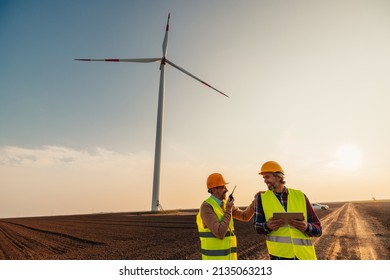 Two Engineers With Safety Helmets Using Walkie Talkie And Digital Tablet While Out Working In A Remote Location Near Wind Turbines