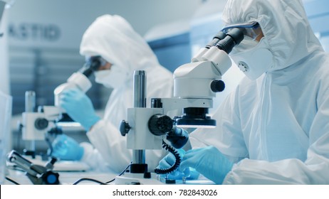 Two Engineers/ Scientists/ Technicians in Sterile Cleanroom Suits Use Microscopes for Component Adjustment and Research. They Work in an Electronic Components Manufacturing Factory.