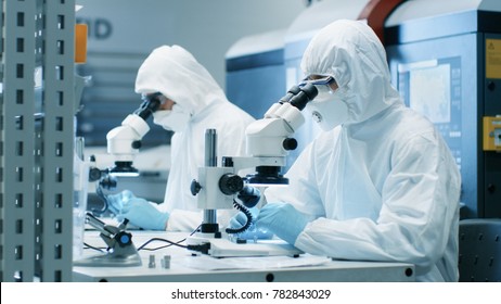 Two Engineers/ Scientists/ Technicians In Sterile Cleanroom Suits Use Microscopes For Component Adjustment And Research. They Work In An Electronic Components Manufacturing Factory.