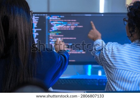 Two engineers seen from behind studying source code on large monitors facing a wall of monitors filled with source code. The candid moment captures modern software development, modernoffice.