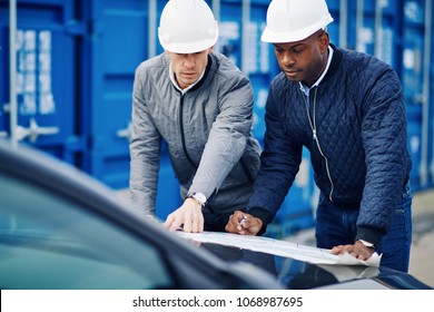 Two engineers standing by freight containers on a commercial shipping dock leaning on the hood of a truck discussing blueprints