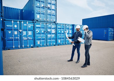 Two engineers standing by freight containers in a commercial shipping yard reading blueprints and talking on a cellphone  