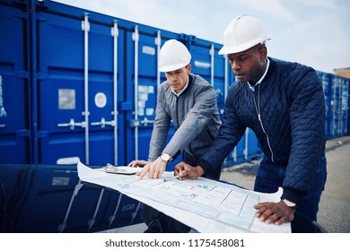 Two engineers standing by freight containers on a commercial shipping dock leaning on the hood of a truck discussing building plans